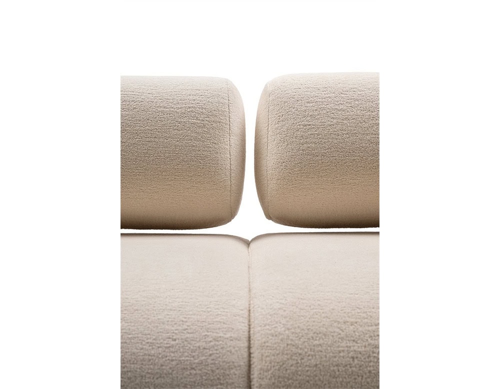 A detailed view of the Evolis Next Sofa showcasing the two cylindrical backrests and the seamless connection of the seat cushions. The fabric has a soft, light tone with a tactile texture.