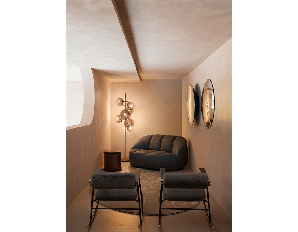 Lou Lou Rock Chair in an intimate setting. Two chairs in dark upholstery face a matching compact sofa, all arranged over a round rug. The space is softly lit by a sculptural floor lamp and features warm neutral tones.