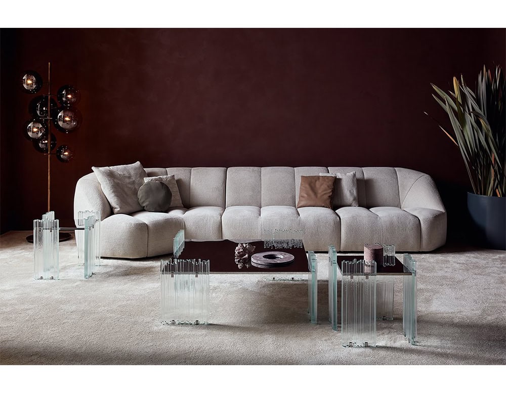 GR Mirage Coffee Table collection arranged in a living room. The clear glass and mirrored tables are placed in front of a soft beige sofa, set against a deep burgundy wall, creating a refined and modern atmosphere.