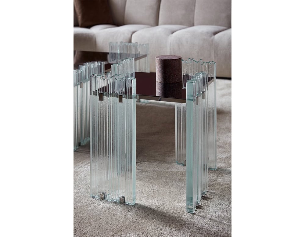 GR Mirage Coffee Table detail in a living room. The clear glass legs and dark reflective top contrast with the soft beige sofa and textured carpet, emphasizing the table's sculptural, crystalline aesthetic.