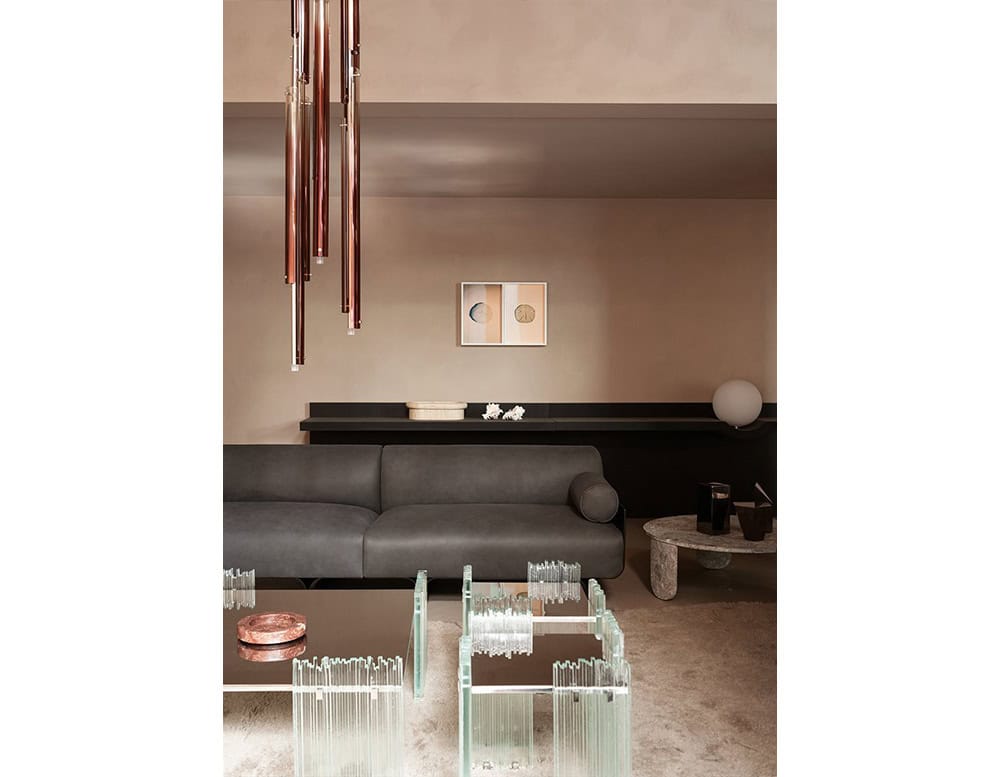 GR Mirage Coffee Table set displayed in a refined living space. The clear glass legs and smoked glass tops harmonize with a charcoal sofa, soft beige carpet, and bronze pendant lighting, creating a modern, elegant atmosphere.