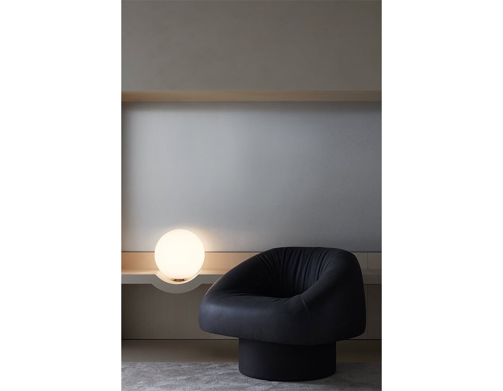 The GR Olivia Chair, upholstered in deep black leather, sits in a calm, softly lit space with neutral walls and a pale console. A spherical white lamp adds a warm glow, enhancing the refined and minimalist atmosphere.
