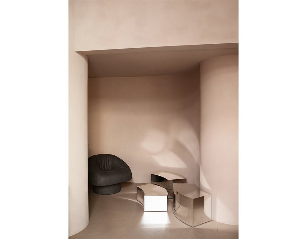 The GR Olivia Chair in charcoal black rests in a curved alcove with soft beige walls. Polished steel tables reflect light, creating a serene, sculptural scene that highlights the contrast between textures.