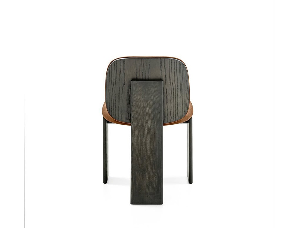 The GR Saki Chair presents a sculptural back view, showcasing its bold central back leg in dark wood with a pronounced grain. The softly rounded backrest wraps in camel leather, highlighting the chair's mix of softness and architectural strength.
