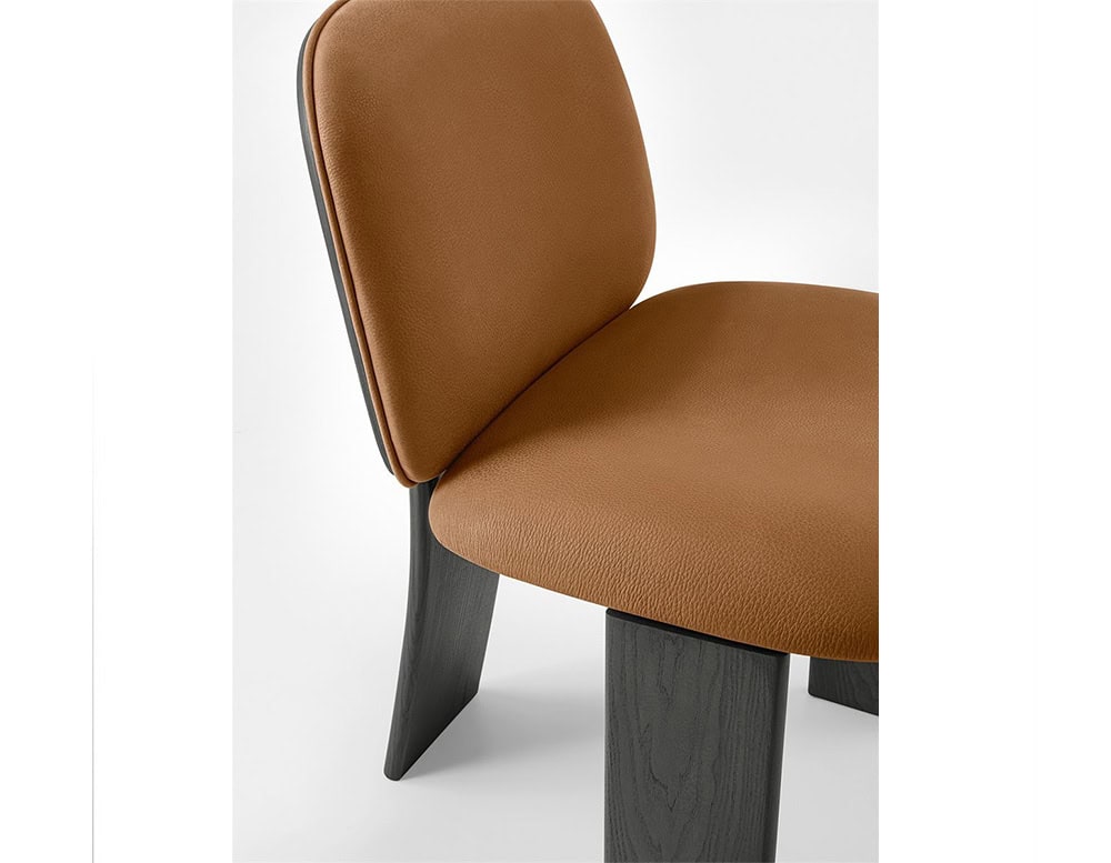 The GR Saki Chair features soft camel leather upholstery with subtle grain texture, paired with dark ash wood legs. The close-up highlights the chair's seamless curves, precise stitching, and the bold contrast between materials.