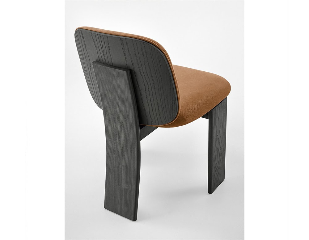 The GR Saki Chair reveals its sculptural back, showcasing dark ash wood with a visible grain and a central curved support. The camel leather seat softly contrasts the bold, architectural lines of the chair's rear profile.