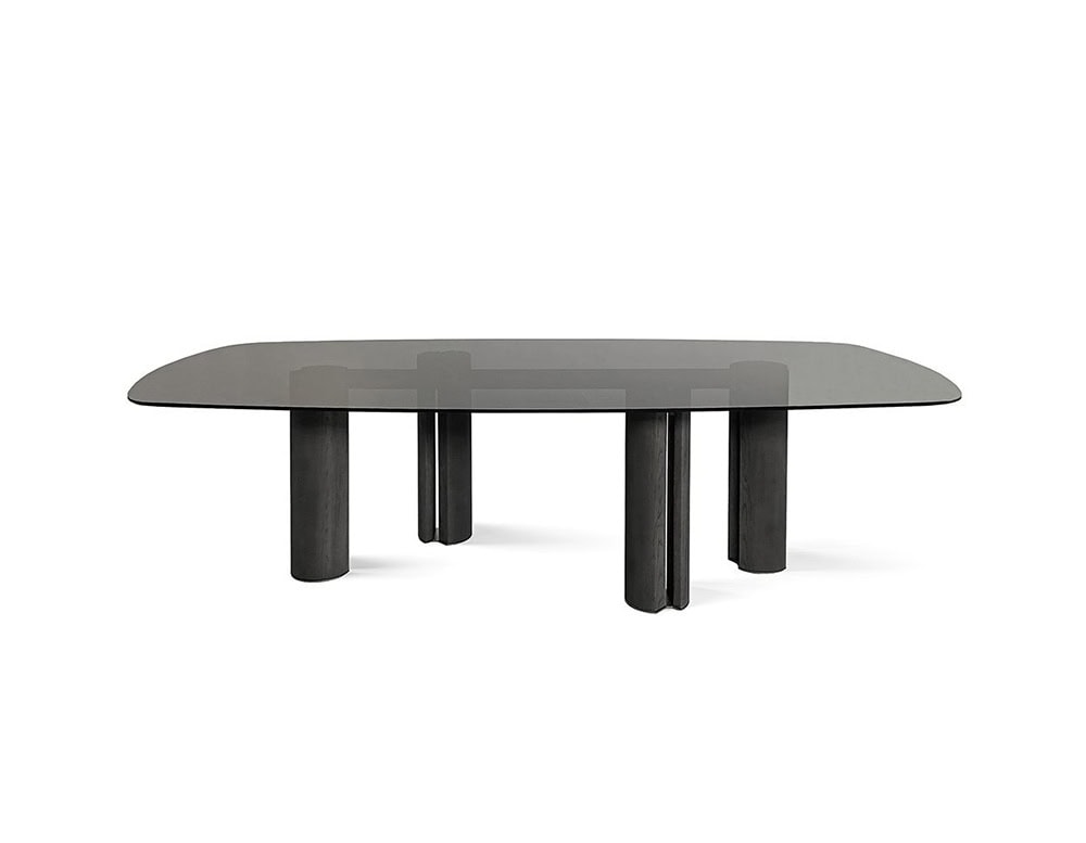 The GR Simpodio W Table features a smoked glass top with softly curved edges. Its charcoal-toned cylindrical legs create a bold, architectural contrast beneath the dark transparent surface.