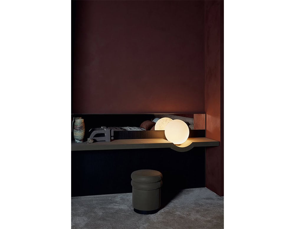 The GR Stresa Mirror anchors a moody corner with its integrated shelf and glowing globe. The composition features deep wood tones, antiqued mirror detailing, and soft lighting, creating a refined and atmospheric vignette.