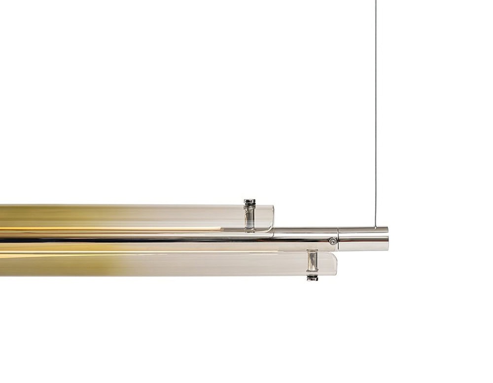 The GR Tratto Suspension Light highlights precision in detail, with its polished linear frame softly transitioning from metallic silver to golden tones, adding a refined amber shimmer to the composition.