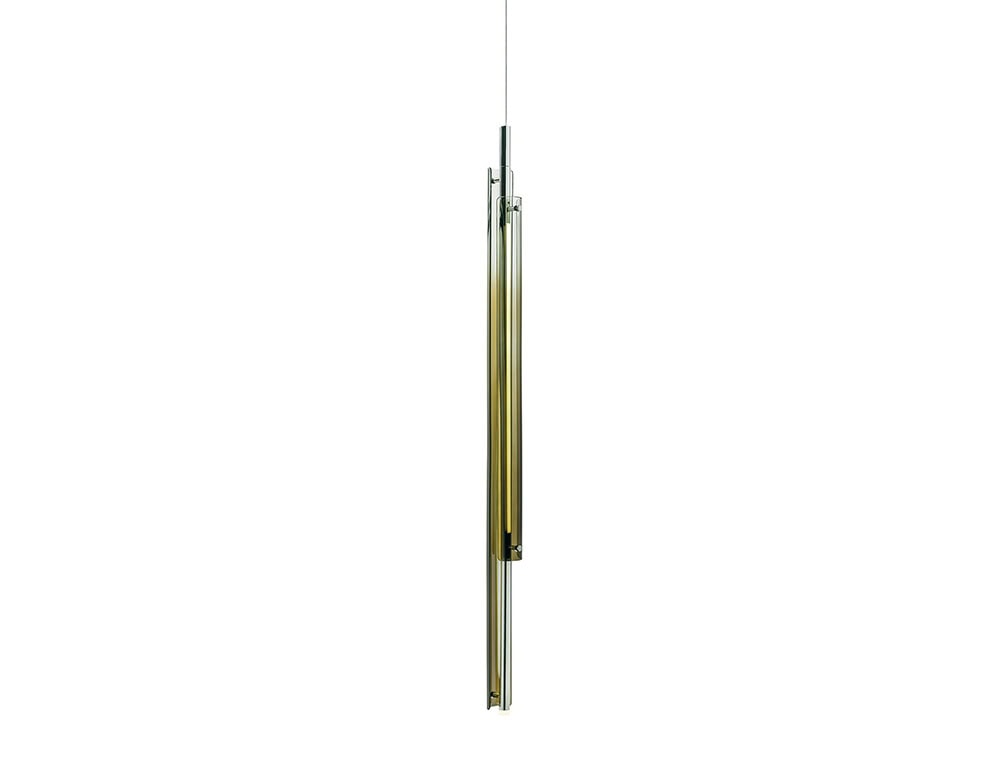The GR Tratto Suspension Light is presented in a vertical composition, highlighting its slender tubular profile. Subtle amber hues blend with polished metal, creating a refined, modern accent.