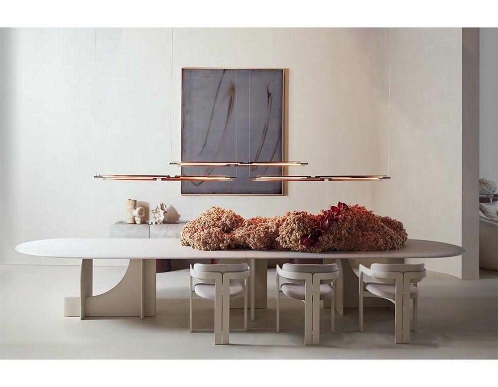The GR Tratto Suspension Light elegantly floats above the table in slim, linear forms. Its warm metallic tones emit a soft, amber glow, adding understated warmth to the neutral dining setting.