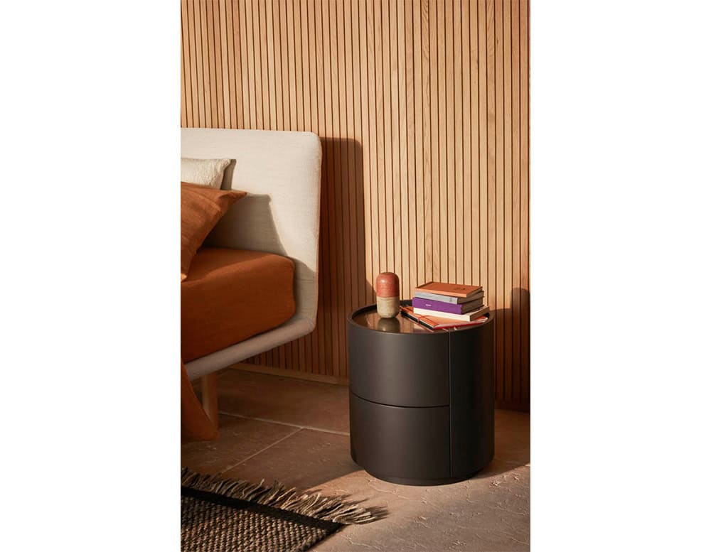 The Pian Dedalo Nightstand features a compact, cylindrical design in matte black with two drawers, adding modern elegance to a cozy bedroom scene with warm wood paneling and soft textiles.