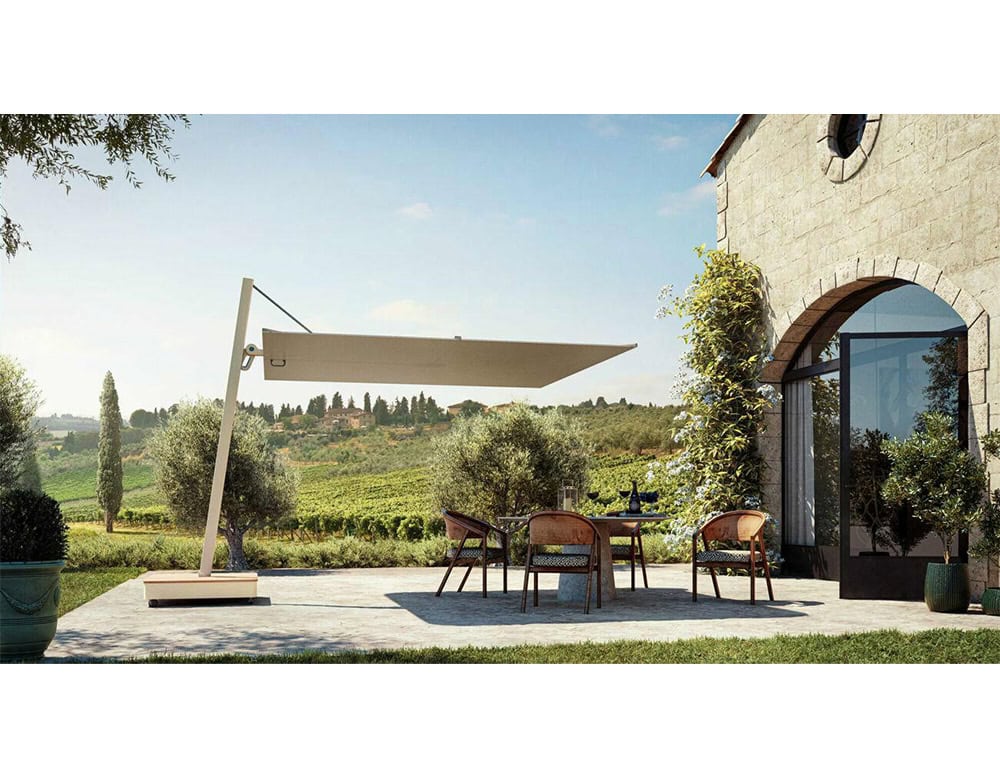 Versa UX Umbrella features a pale beige canopy and angular cantilever arm, casting sculptural shade over a rustic patio. Surrounded by vineyard hills and olive trees, the scene blends modern design with serene countryside charm.