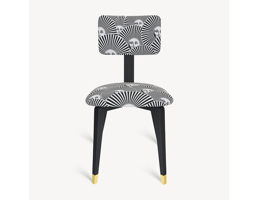 The Fornasetti Soli a Ventaglio Chair features a bold black and white radial design with iconic sun faces. Its tapered black legs are finished with brass-capped feet for an elegant accent.