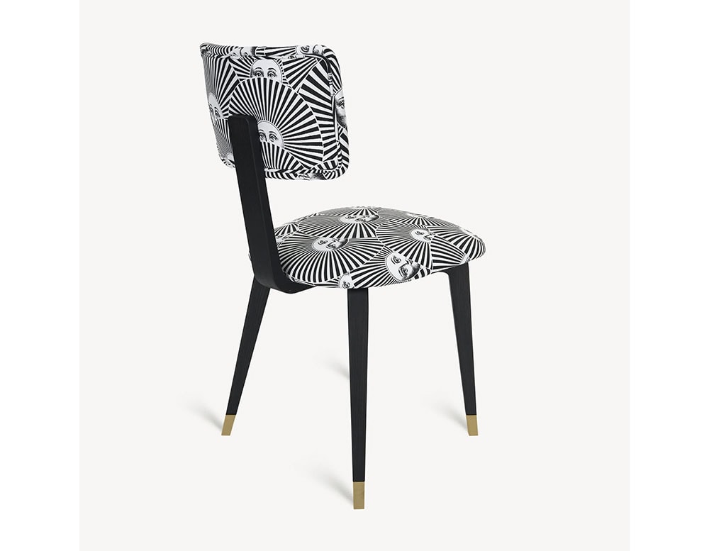 The Fornasetti Soli a Ventaglio Chair combines whimsical sun faces with striking radial lines, creating a bold contrast against its sleek black wood frame and brass-detailed legs.