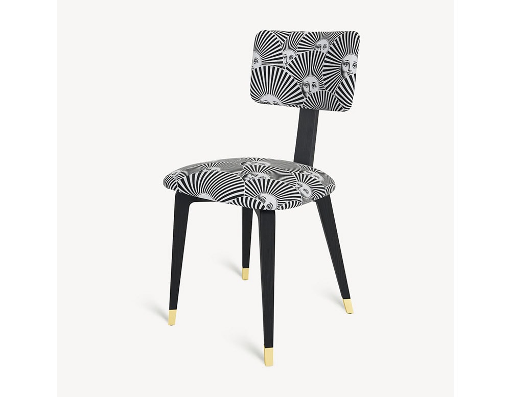 The Fornasetti Soli a Ventaglio Chair features radiant sun faces interlaced with black and white graphic rays, set on a refined black frame with brass-tipped legs for a playful yet sophisticated statement.