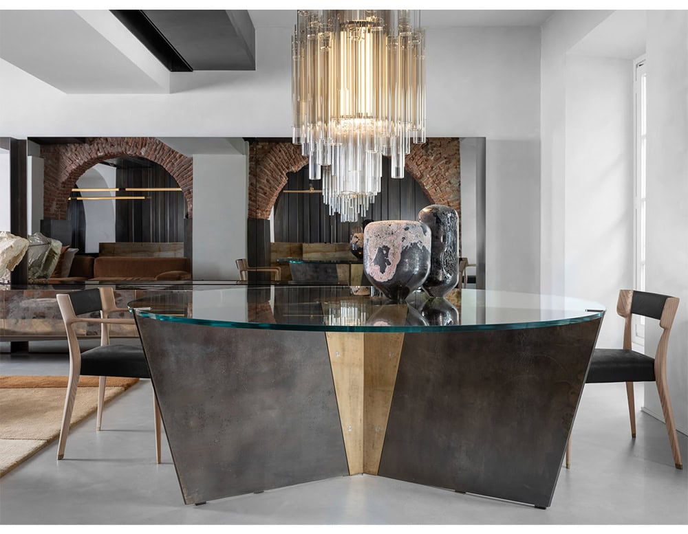 The Hen Chair VA features a sculpted backrest and pale wood frame with black upholstered seat, blending elegance and contemporary design around a striking glass and metal dining table.