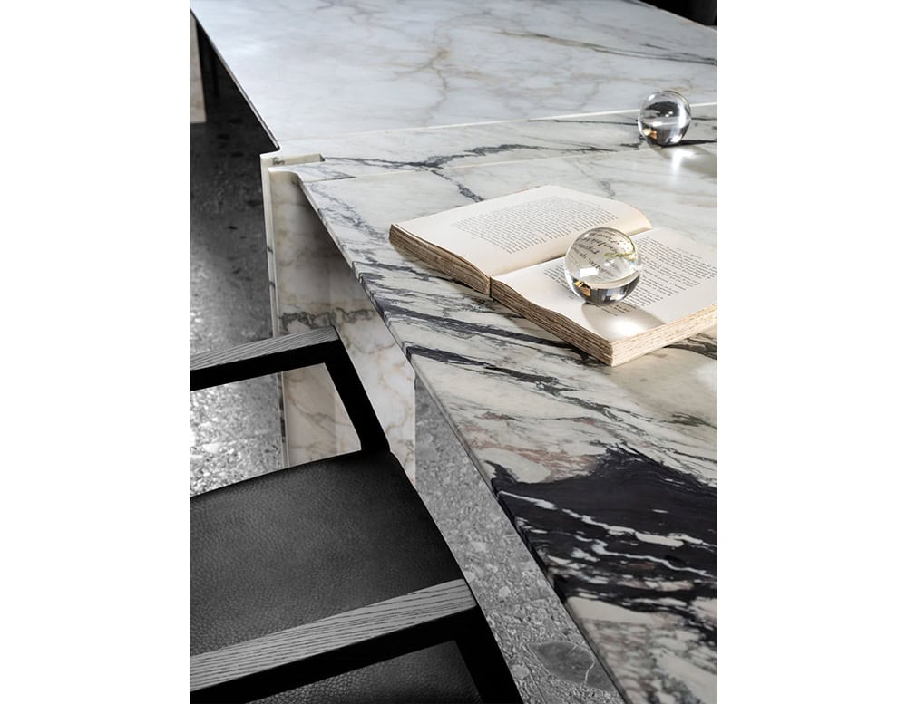 A close up of the Hen Mantis Chair reveals its refined craftsmanship. Black leather upholstery meets a sharply angular wooden armrest in a striking architectural profile. Shown beside a bold marble table.