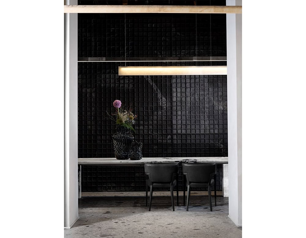 Hen Mantis Chairs with low profile and curved backrests pair with a marble dining table. The chairs' matte black finish complements the striking black tile wall and sculptural ceramic centerpiece, grounding the space with modern elegance.