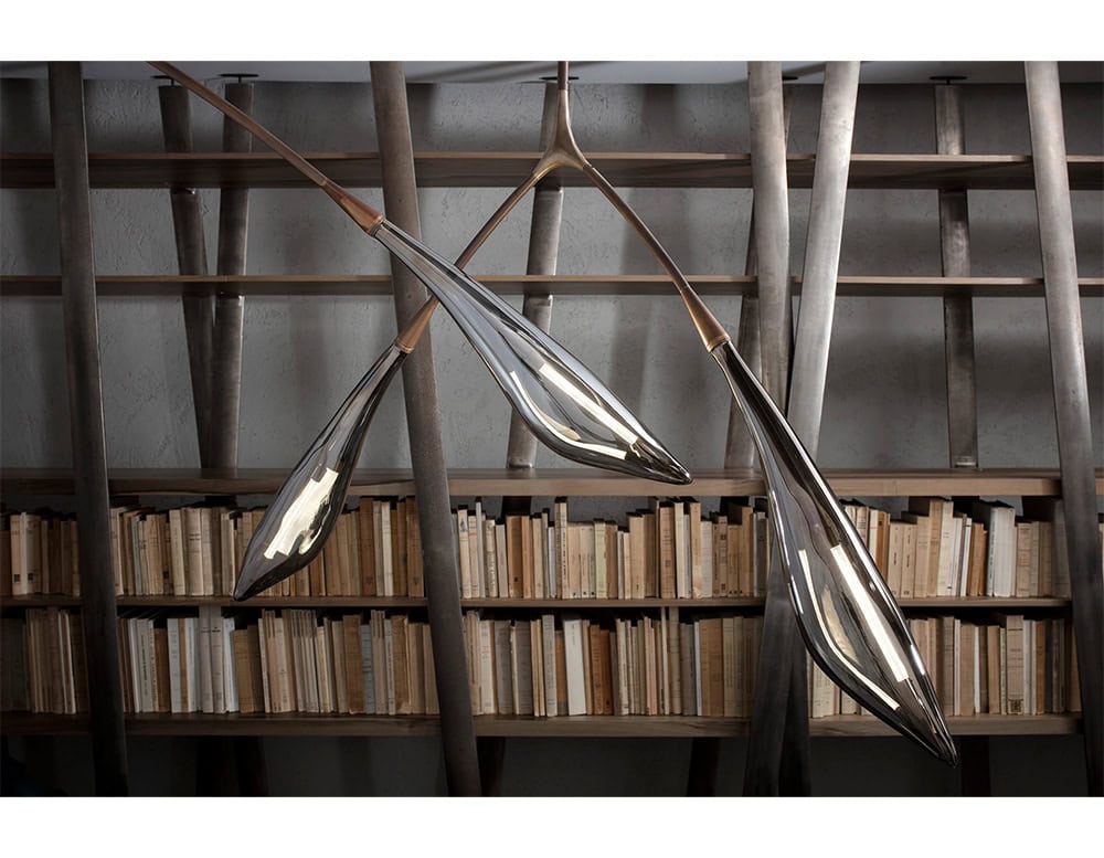 The Hen Maple ceiling light features elegant bronze arms and glass shades resembling leaves. Suspended in front of a modern bookshelf, it blends organic form with architectural sophistication.