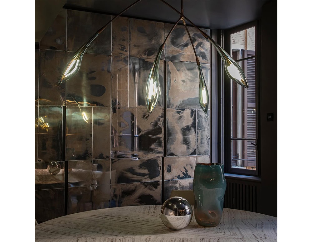 Hen Maple ceiling light floats above a sculptural table, its fluid glass diffusers casting soft glows against mirrored wall panels. The piece adds organic elegance and ambient warmth to a richly textured, contemporary dining setting.