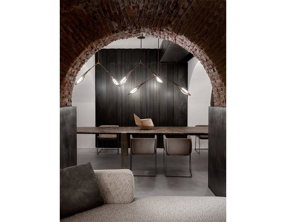 Hen Maple ceiling light arcs elegantly over a long dining table, blending organic form with warm illumination. Set beneath a brick archway, it contrasts beautifully with the dark paneled wall and soft neutral furnishings.