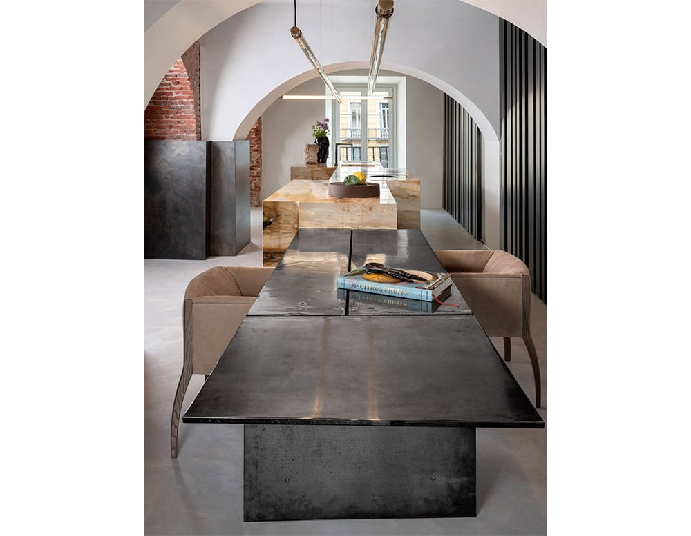The Hen Shelly Chair features a cocooned seat with tactile upholstery and gently tapered wood legs. Its soft lines and inviting form complement the raw texture of the metal dining table and the architectural details of the space.