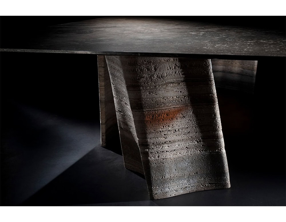A sculptural base with an organic texture supports the dark tabletop. The form evokes carved stone with layers and folds that play with shadow and light for a commanding earthy presence.