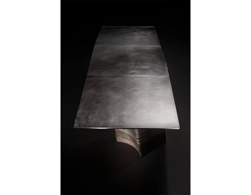 A dramatic rectangular top with a velvety matte finish extends across a monolithic base. The soft gradient of dark tones on the surface enhances the piece’s moody, sculptural character.