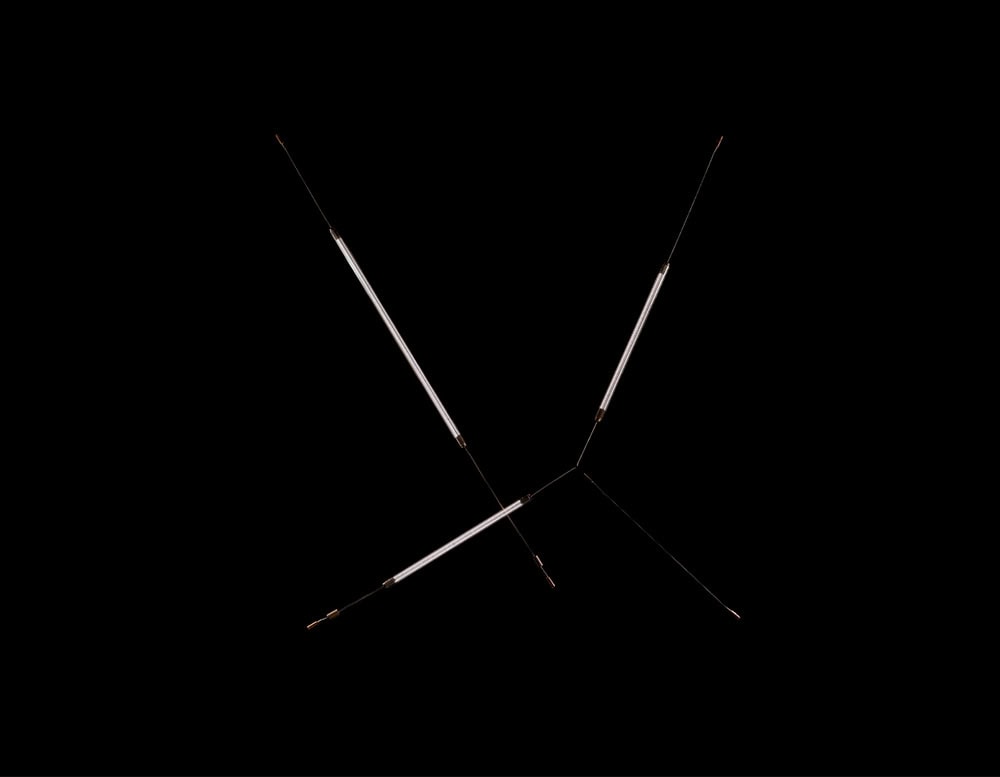 Hen Spyder wall light features a dynamic, linear form with intersecting arms and slim illuminated tubes. The minimal structure evokes a sense of motion and precision, standing out dramatically against the black background.