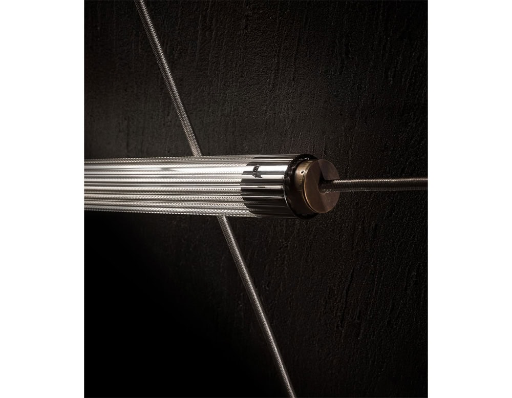Detail of the Hen Spyder wall light reveals its ribbed glass tube suspended by taut metal cables and anchored with bronze-toned hardware. The close-up highlights its refined, industrial aesthetic and technical precision.