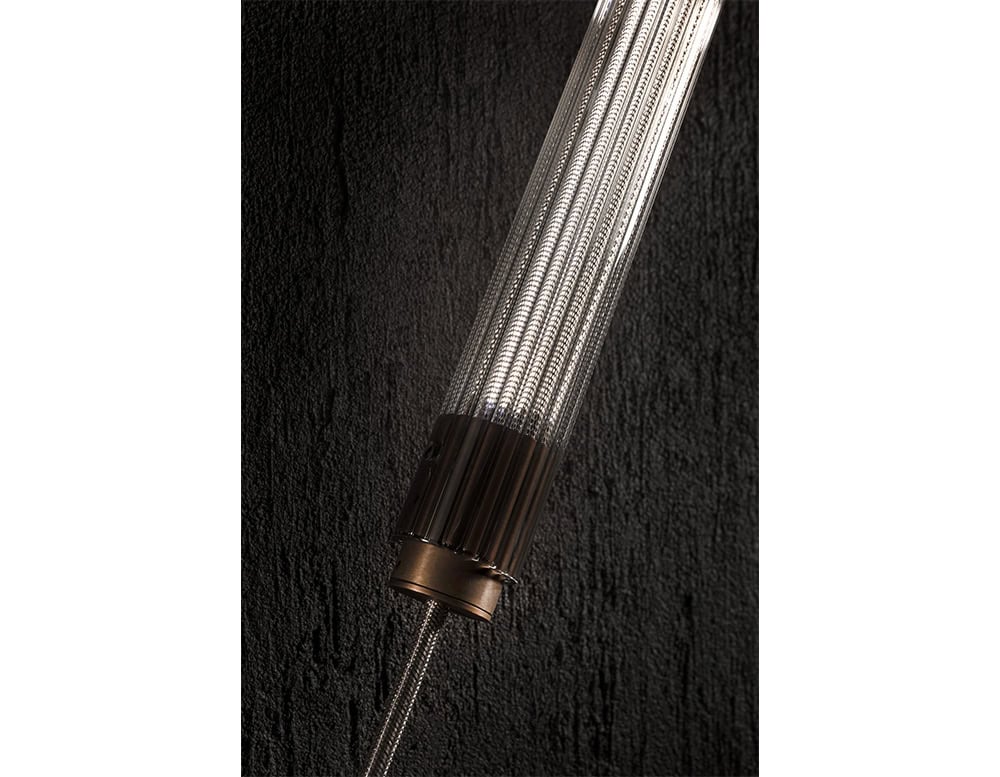 The Hen Spyder wall light’s illuminated ribbed glass tube is anchored with bronze-finished hardware and suspended by braided metal cables, showcasing refined industrial elegance and precise craftsmanship.
