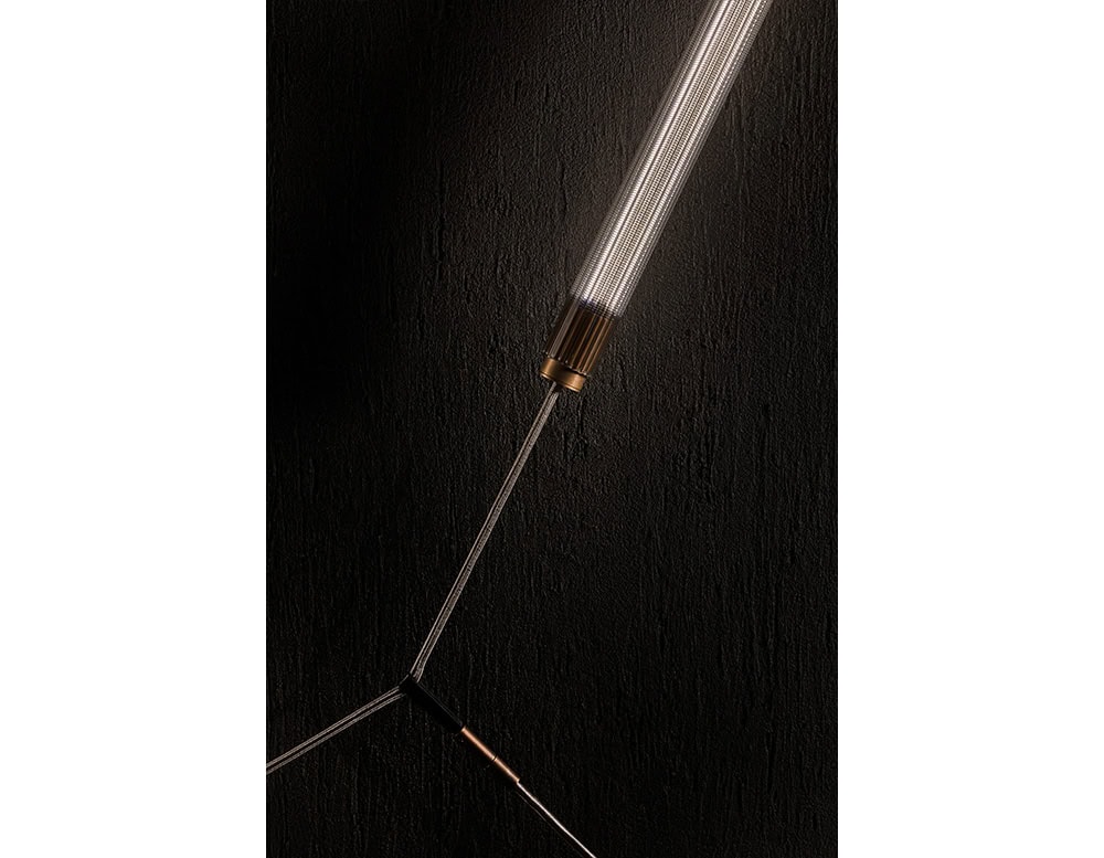 The Hen Spyder wall light reveals a linear ribbed glass diffuser suspended by sleek braided cables and capped with bronze-toned accents, delivering a minimalist yet striking aesthetic for modern interiors.