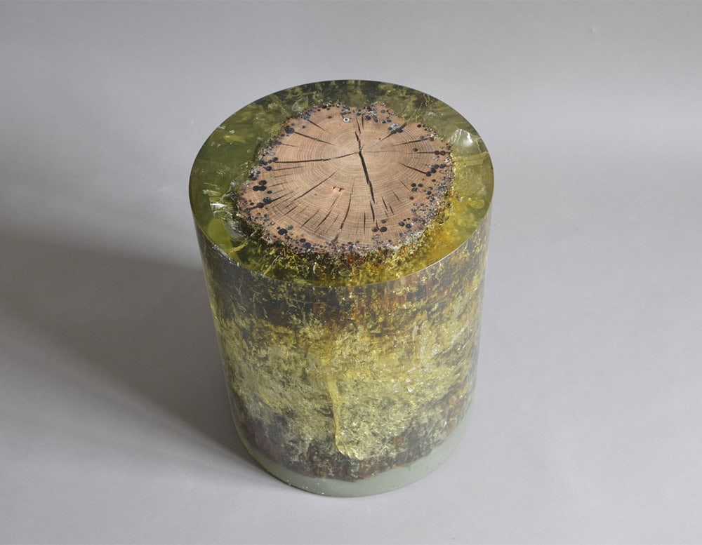 A single cylindrical stool encases a cross-section of natural wood in thick, amber-toned resin. Cracks, dark inclusions and trapped air bubbles add organic texture and raw visual depth.