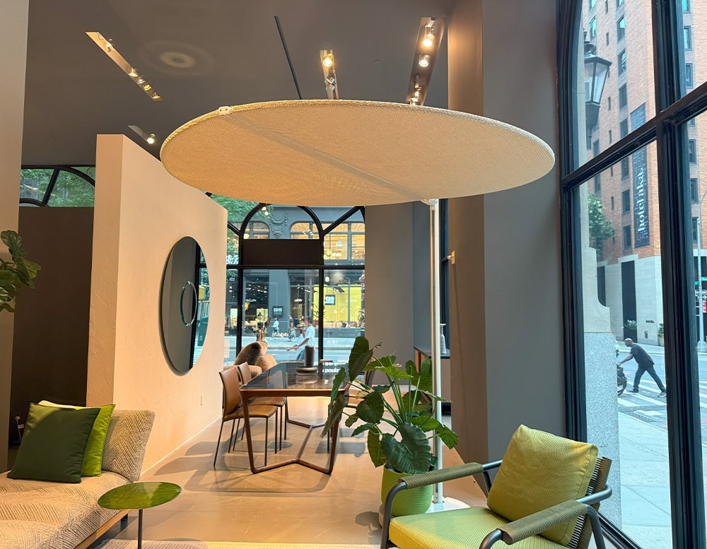 Minimalist round umbrella with a light woven canopy and slim white stand. Provides stylish shade beside a lounge chair in a modern showroom with tall arched windows.