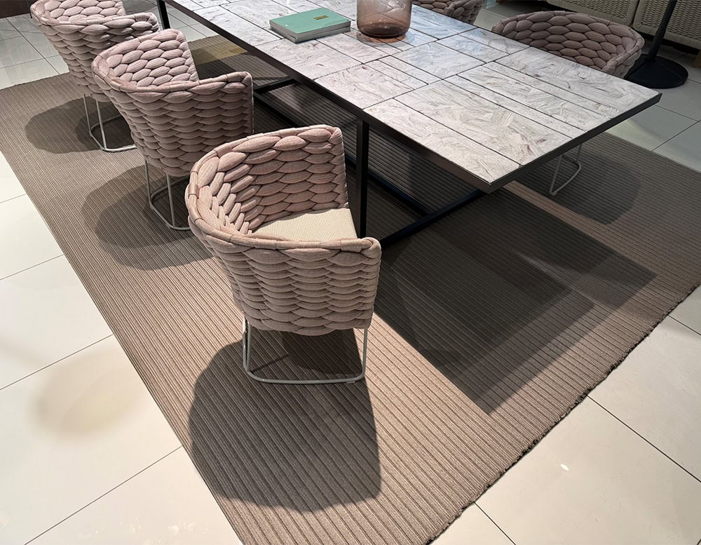 Plush woven armchairs surround a sleek tile top table, set over a ribbed neutral rug that enhances texture and comfort for outdoor dining spaces.