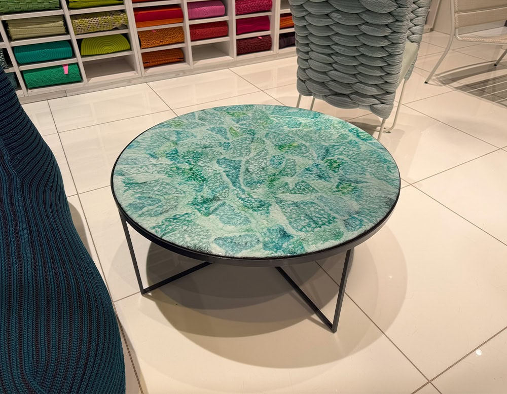 Round coffee table with an artistic blue-green glass top and slim black metal base. Vivid, ocean-inspired tones create a striking visual centerpiece.