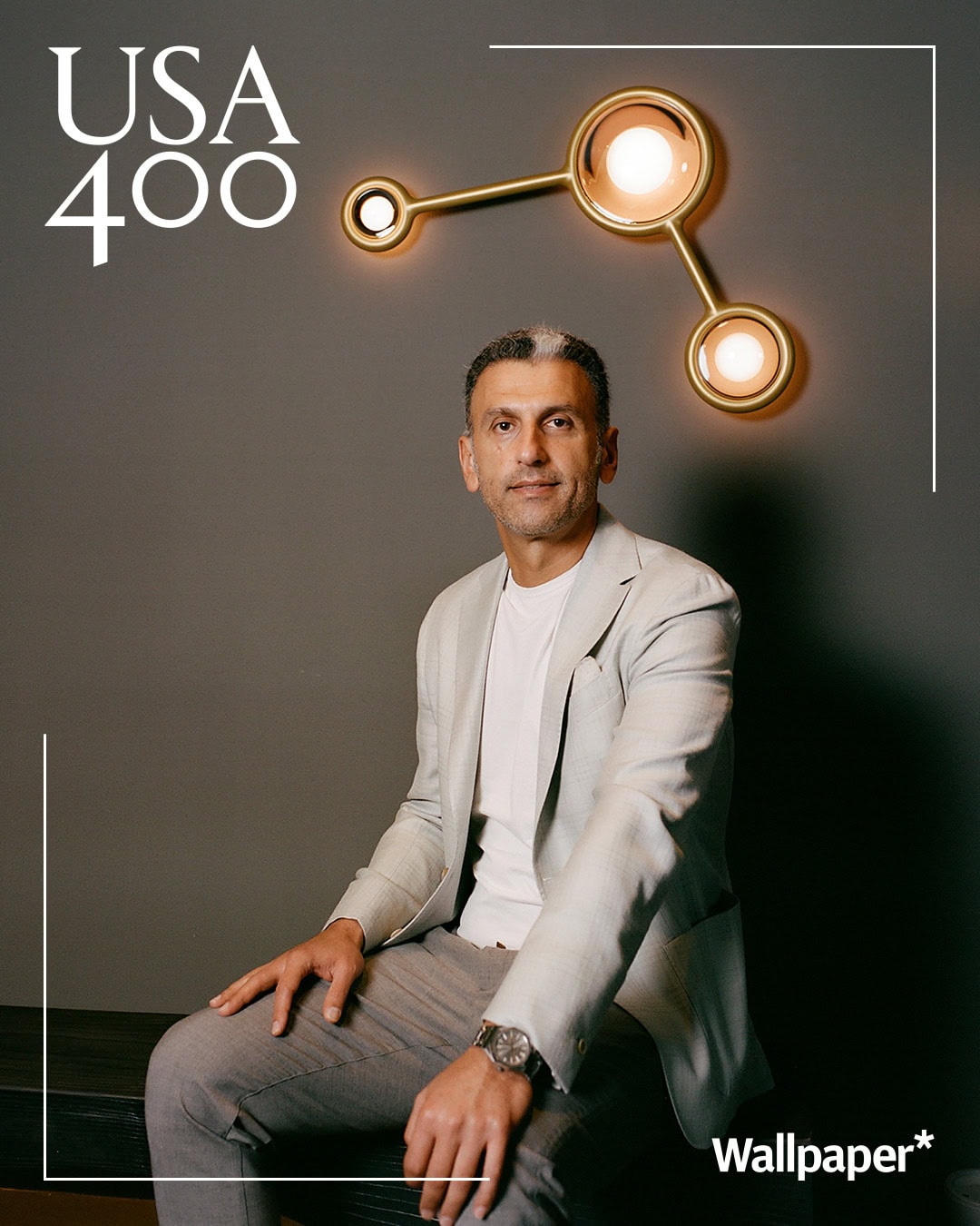 A confident portrait featuring a man seated against a warm gray backdrop with sculptural lighting above him. His relaxed posture and thoughtful expression create a refined and modern atmosphere perfect for an editorial setting.