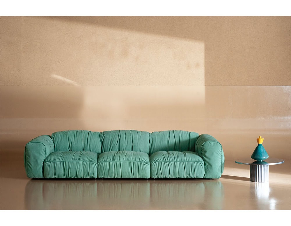 Green upholstered sofa with four wide seats and gathered fabric detailing, set against a beige wall with reflective flooring and a modern round side table holding a sculptural vase.