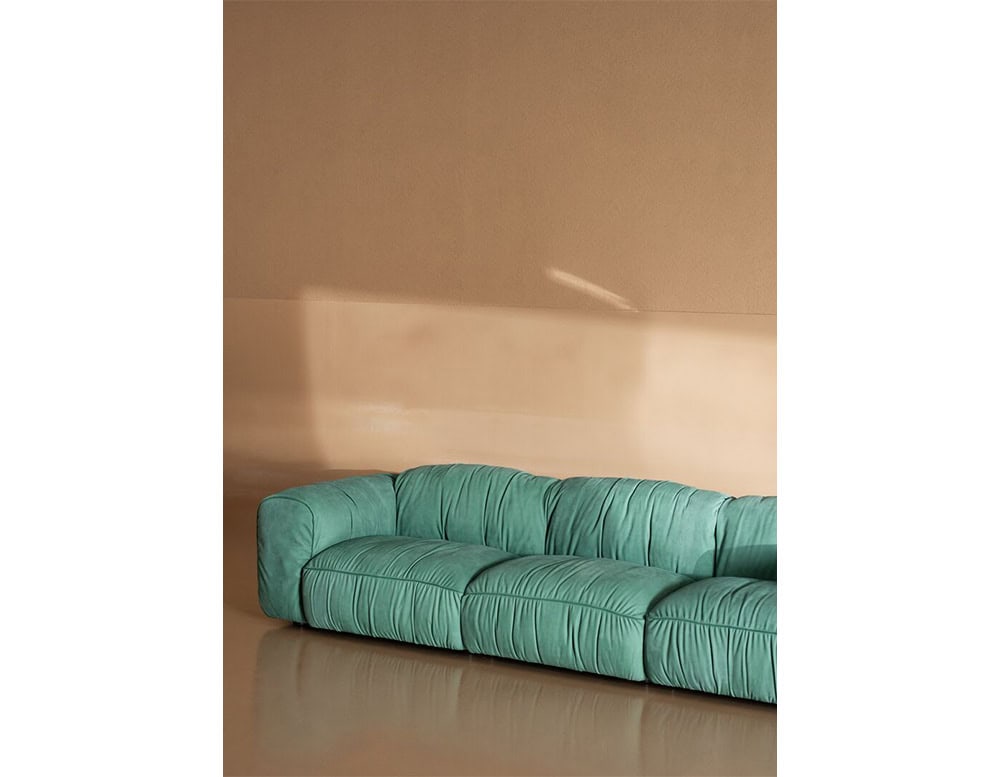 Green upholstered sofa with low deep seating and gathered fabric texture, placed in a minimalist room with neutral beige walls and a reflective floor.