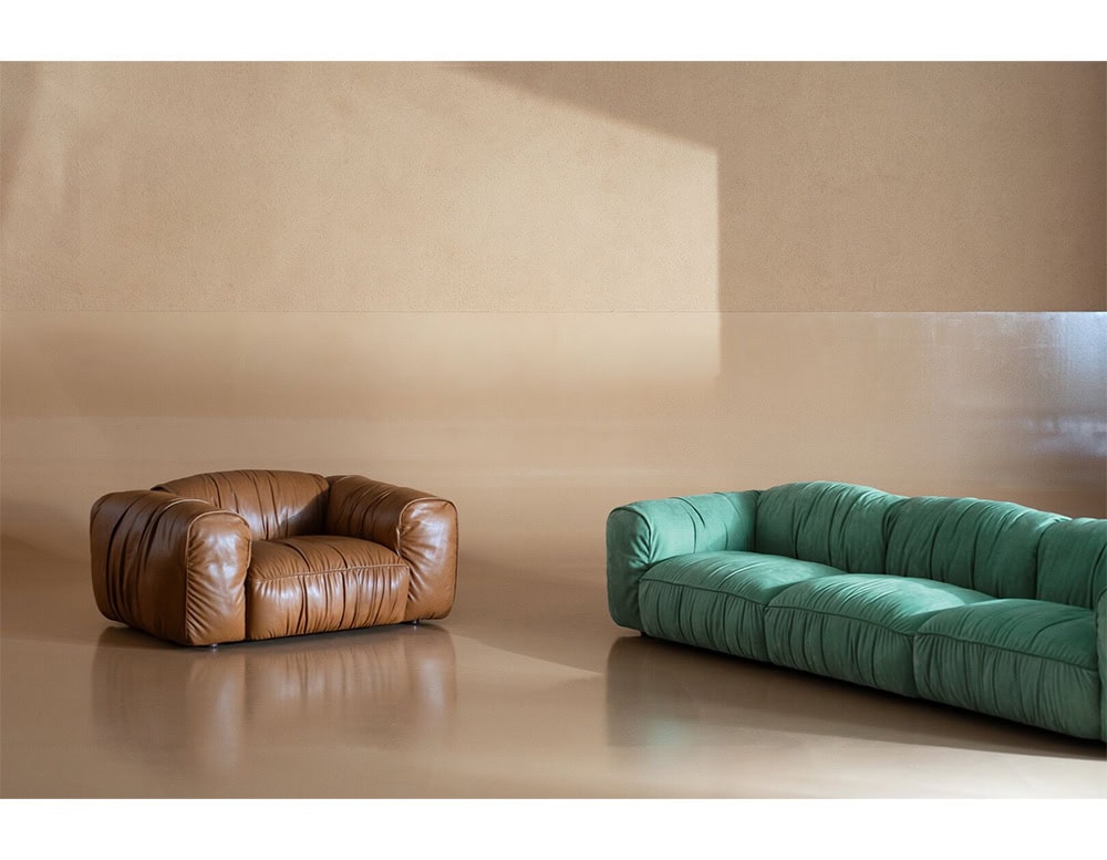 Contemporary brown armchair and green sofa with plush, ruched fabric upholstery sit in a minimalist room, their textures highlighted by sunlight reflecting on the smooth floor.