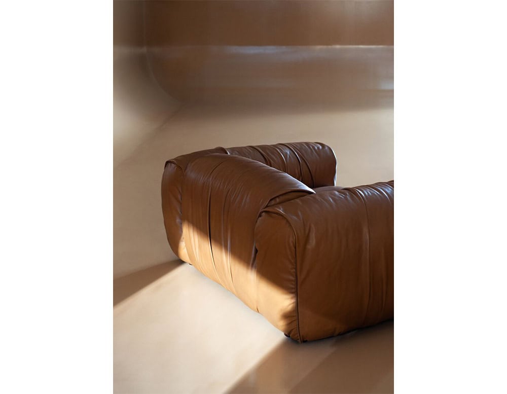 A low profile brown leather armchair with deep cushions is placed in a minimalist room, highlighted by soft natural light that enhances its smooth texture and modern appeal.