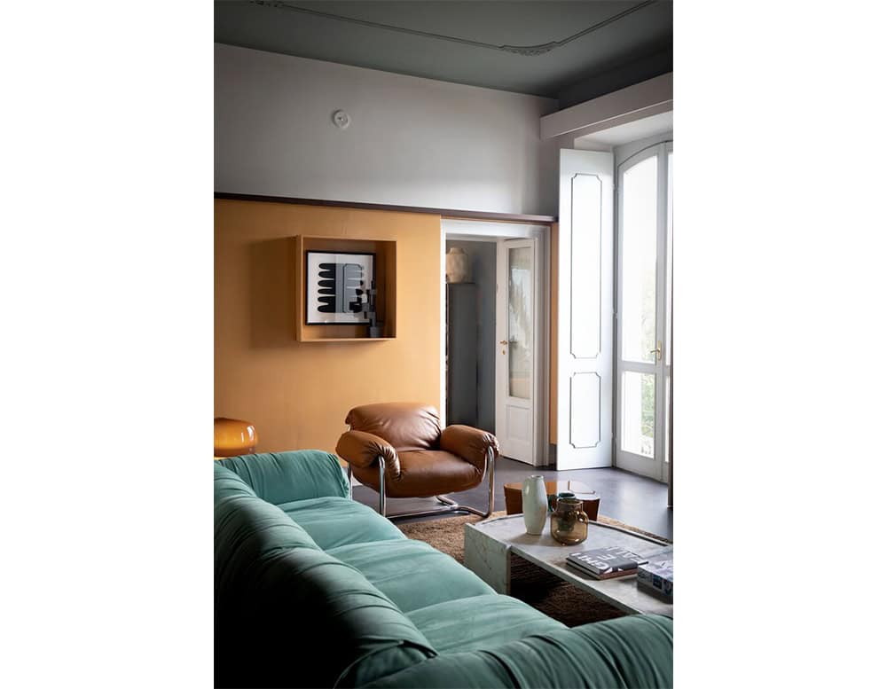 Living room with a green sofa and a brown leather armchair arranged around a low marble coffee table, styled with decorative vases and books near tall windows framed by white shutters.