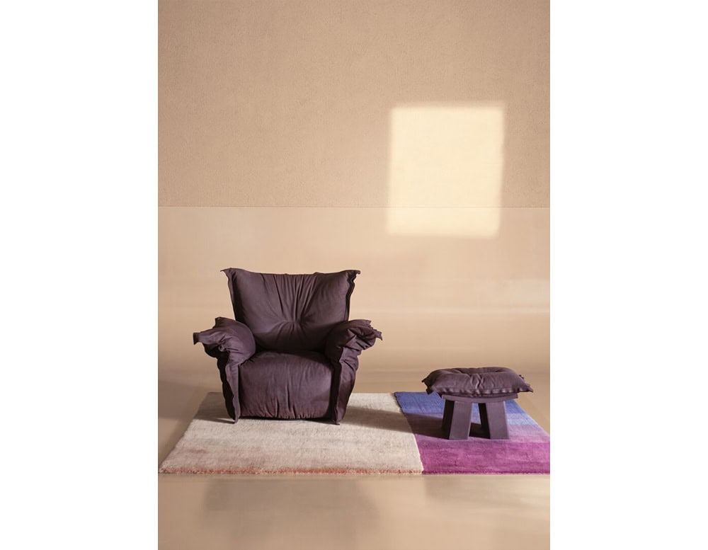 Dark purple armchair with matching cushioned footstool placed on a color-blocked rug in soft neutral surroundings, creating a stylish and comfortable reading or lounge corner.