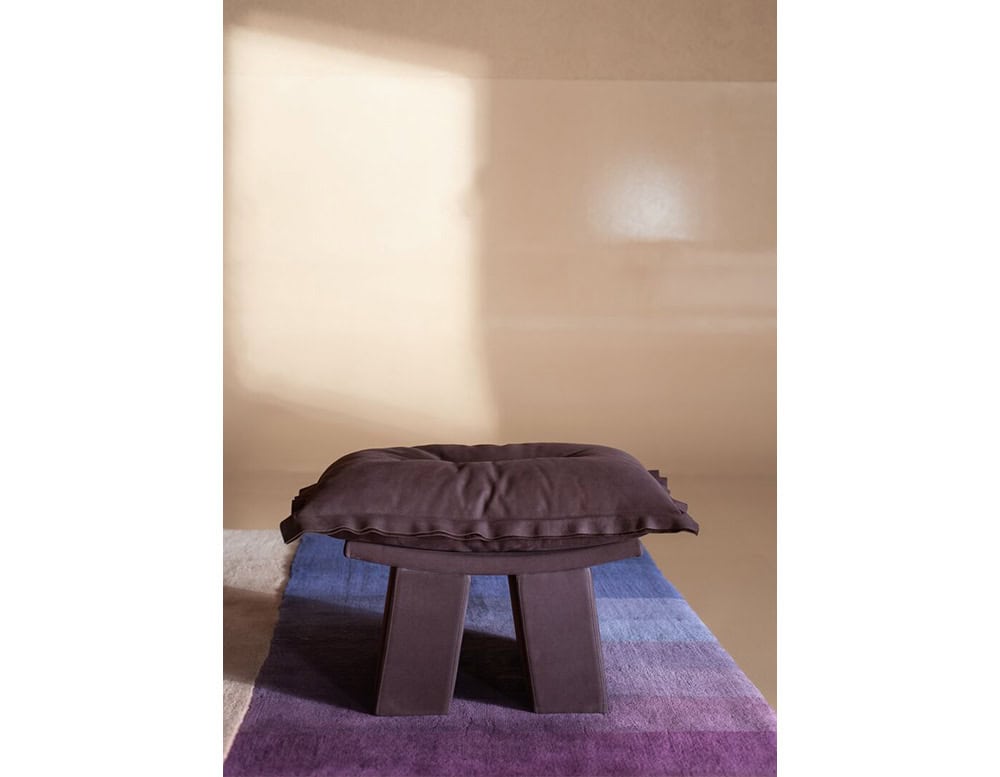 Stylish footrest featuring a plush purple cushion on a solid base, set on a soft rug with gradient tones of blue and purple for a modern and coordinated look.