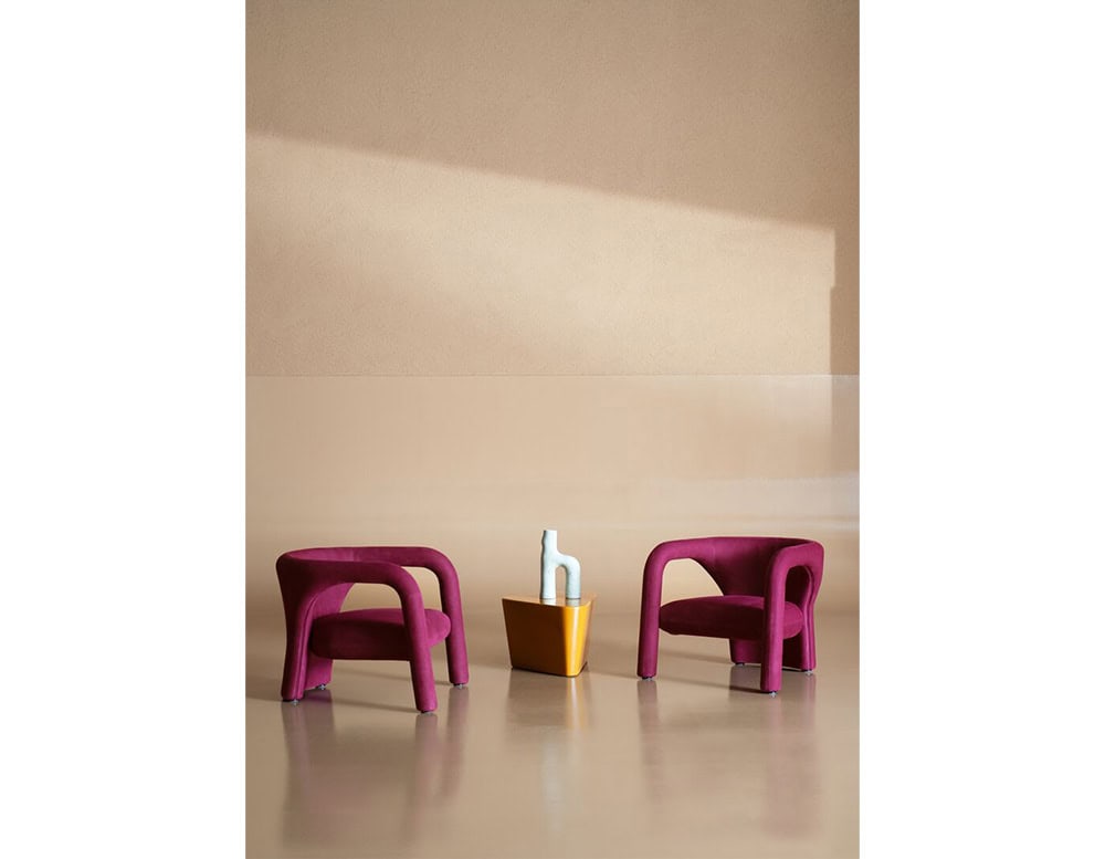 Pair of plum-colored armchairs with curved silhouettes and plush seats frame a mustard side table with a sculptural white vase, blending geometric contrast and modern elegance in a soft neutral space.