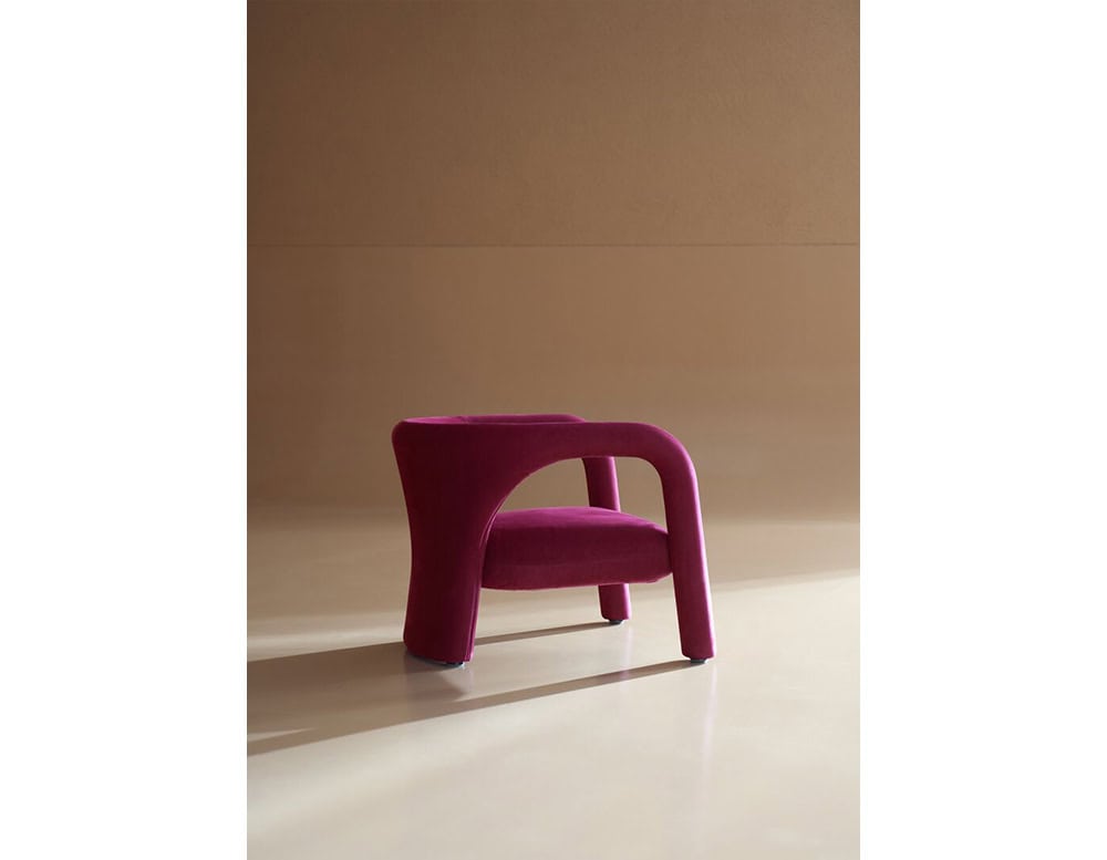 A sculptural armchair in bold magenta velvet, defined by fluid, rounded lines and an embracing form that balances modern artistry with plush comfort.