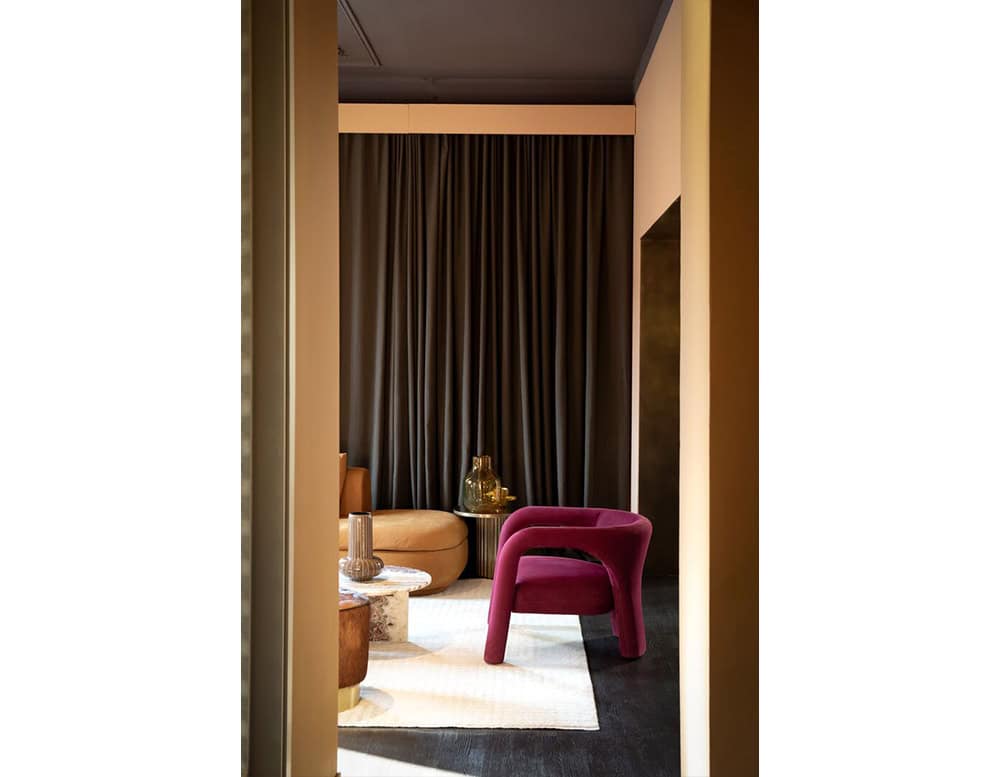 A magenta Aimee Armchair complements the earthy palette of tan seating, marble tables, and dark drapery, creating a refined balance of modern elegance and inviting comfort.