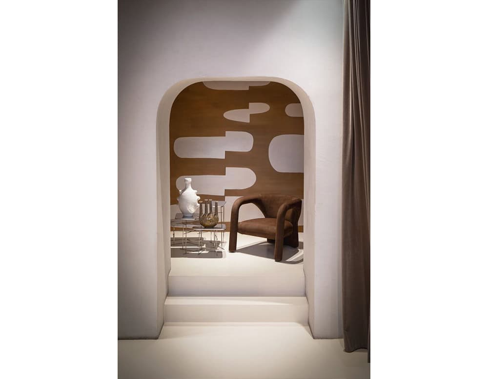 A sculptural brown armchair with fluid, continuous lines and plush upholstery sits within an arched alcove. Paired with ceramic vases and a metal side table, it forms a serene, modern vignette in soft neutral tones.