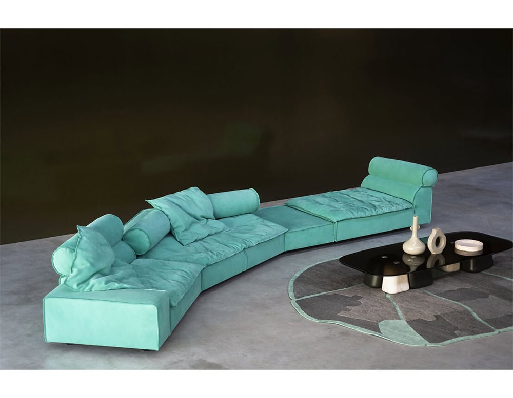 A modular sofa in vivid turquoise fabric with plush cushions and cylindrical backrests that offer relaxed comfort, paired with a sculptural coffee table and geometric rug for a bold yet refined modern setting.